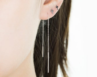 May include: A close-up of a person's ear adorned with three silver cross earrings. Two small cross studs are visible, along with a pair of chain earrings with a vertical bar pendant. The earrings are approximately 4.3 inches long.