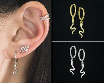 Puede incluir: Pendientes con motivos de serpiente en oro y plata. Los pendientes dorados tienen una serpiente colgante y una pequeña gema. Los pendientes plateados tienen un diseño similar. La imagen también muestra una oreja con un pendiente de serpiente y un brazalete de oreja de diamantes.