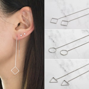 May include: A person's ear with three small stud earrings and a pair of silver threader earrings with a square geometric design. Other earrings with circle and triangle designs are also shown.