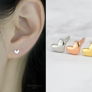 May include: Three heart-shaped stud earrings in silver, rose gold, and gold. One earring is shown worn on an ear. The earrings are a simple design, suitable for everyday wear. The brand name "Bambi&dew" is visible at the bottom.