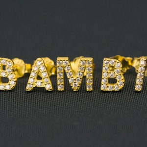 May include: Gold stud earrings spelling "BAMBI" in capital letters. Each letter is set with small, clear, sparkling stones. The earrings are presented on a dark grey textured surface.