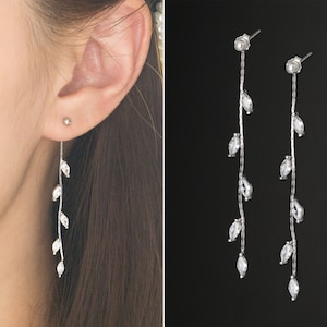 May include: Silver drop earrings with a vine-like design. Each earring has a slender, textured stem with several clear, marquise-cut crystals. The earrings are shown on a person's ear and against a dark background.