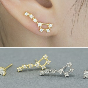 May include: Gold and silver constellation earrings featuring clear cubic zirconia stones. The earrings are designed as a constellation, with a stud and a climber earring. The climber earring is worn along the ear, with the stud earring on the lobe.