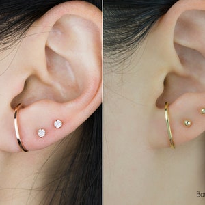 May include: Two images of ears displaying jewellery. One ear has a rose gold hoop earring and two small diamond stud earrings. The other ear features a gold hoop earring and two small gold ball stud earrings.