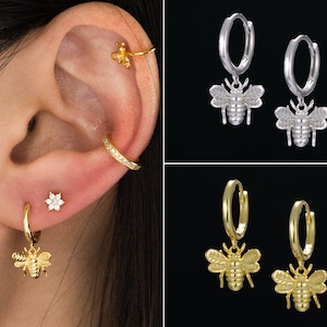 May include: Earrings with bee and flower designs. The image shows gold and silver hoop earrings with bee pendants, and a small flower stud earring. The earrings are displayed on an ear.