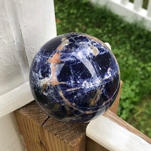 May include: A polished sodalite sphere, displaying a deep blue color with veins of white and orange. The gemstone ball is resting on a wooden surface, with a white fence and greenery in the background.