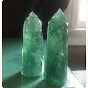 Puede incluir: Dos puntas de cristal de fluorita verde, también conocidas como "amatista verde", se muestran sobre una superficie verde. Los cristales son translúcidos y tienen una textura natural y rugosa.