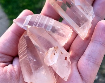 May include: A collection of four clear quartz crystals with natural striations and a slight pink hue. The crystals are raw and unpolished, with sharp points and edges.
