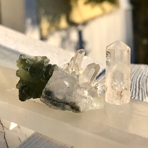 Pode incluir: Três cristais: uma pedra texturizada verde escuro, um aglomerado de cristais de quartzo transparente e um cristal transparente e pontiagudo. Os cristais são exibidos em uma superfície branca, com um brilho suave da luz solar.