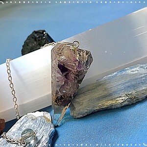 May include: A purple amethyst crystal pendulum with a silver chain and a clear quartz crystal base. The pendulum is on a blue surface with other crystals.