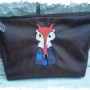 Könnte beinhalten: Eine braune Tasche aus Kunstleder mit Reißverschluss, auf der ein Fuchs mit einem blau-schwarzen karierten Hemd und einer Monokel gestickt ist.