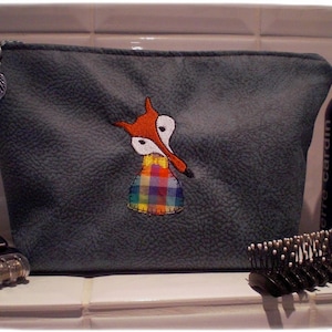 Könnte beinhalten: Eine graue Reißverschlusstasche mit einem aufgestickten Fuchs auf der Vorderseite. Der Fuchs trägt ein kariertes Hemd und hat eine rote Nase und orangefarbene Ohren. Die Tasche liegt auf einer weißen Oberfläche.