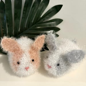 May include: Two fluffy knitted bunny toys. One bunny is white with pink ears and a pink nose. The other bunny is white with gray ears and a gray nose.
