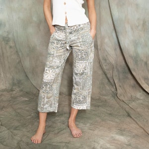 Op de afbeelding: Een vrouw draagt een wit, mouwloos gebreid topje met knoopsluiting en een broek met patroon. De broek heeft een geometrisch ontwerp in tinten bruin en groen.