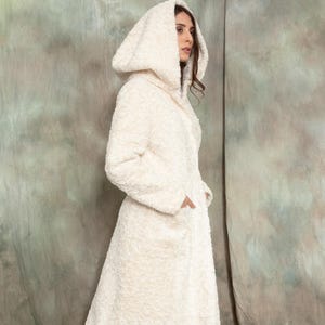 Könnte beinhalten: Langer, cremefarbener Kapuzenmantel mit strukturiertem, plüschigem Aussehen. Der Mantel hat ein durchgehendes Design, Taschen und wird mit schwarzen Stiefeln getragen. Der Gesamtstil deutet auf Wärme und Komfort hin.