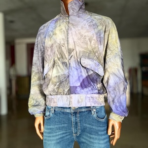 Könnte beinhalten: Eine helllila und graue Batikjacke mit Reißverschluss und zwei Taschen. Die Jacke hat einen Stehkragen und einen gerippten Bund.