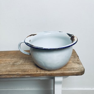 Può includere: Pentola da camera in smalto bianco con un bordo blu e macchie di ruggine. La pentola è appoggiata su una superficie di legno.