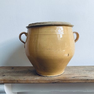 May include: An antique yellow ceramic pot with a lid and two handles. The pot has a rounded body and a flared base. The lid is a lighter yellow. The pot is on a wooden surface. The pot's height is approximately 30 cm.
