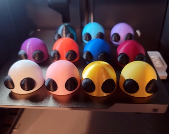 May include: A collection of 12 small, 3D printed alien figurines in various colors, including blue, red, yellow, pink, and purple. Each figurine has two black eyes and a rounded head.
