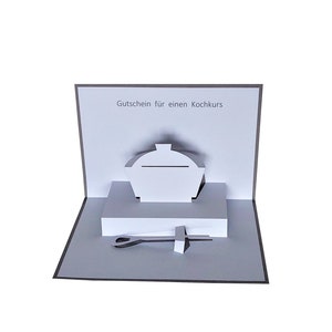 Könnte beinhalten: Eine weiße Pop-up-Karte mit einem 3D-Papier-Ausschnitt eines Kochtopfs mit Deckel und einem Servierbesteck. Der Text "Gutschein für einen Kochkurs" ist oben auf der Karte zu sehen. Die Karte liegt offen auf einer grauen Oberfläche.