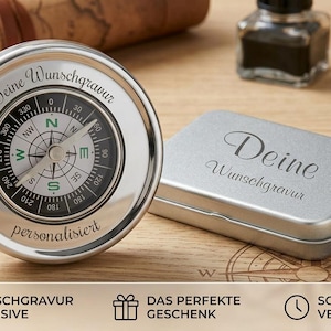 Puede incluir: Una brújula circular de tono plateado con una esfera negra que muestra los puntos cardinales y marcas de grados. La brújula tiene grabados los textos 'Deine Wunschgravur' y 'personalisiert'. Incluye una caja rectangular de metal plateado.