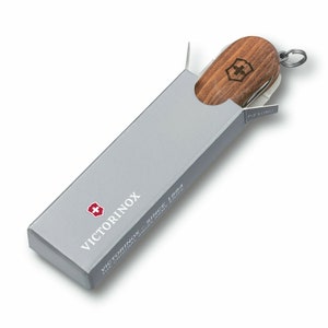 Puede incluir: Una navaja suiza con mango de madera es parcialmente visible dentro de una caja rectangular gris. La caja tiene el logotipo y el texto de Victorinox. La navaja tiene un llavero plateado y se ve una peque&ntilde;a hoja.