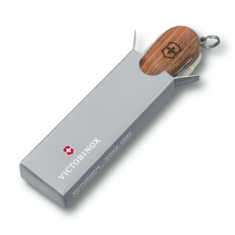 Puede incluir: Una navaja suiza con mango de madera es parcialmente visible dentro de una caja rectangular gris. La caja tiene el logotipo y el texto de Victorinox. La navaja tiene un llavero plateado y se ve una peque&ntilde;a hoja.