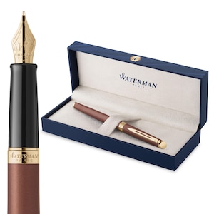 Könnte beinhalten: Ein brauner und goldener Waterman-Stift mit goldener Feder, präsentiert in einer blauen Geschenkbox. Der Stift hat ein schlankes Design mit schwarzem Griff und goldenen Akzenten. Die Box ist mit cremefarbenem Stoff ausgekleidet und trägt das Waterman Paris Logo.