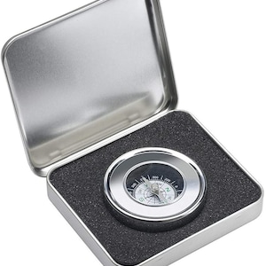 Puede incluir: Una br&uacute;jula plateada con una cara negra y marcas blancas. La br&uacute;jula est&aacute; en una caja de metal plateada con un forro de espuma negra.