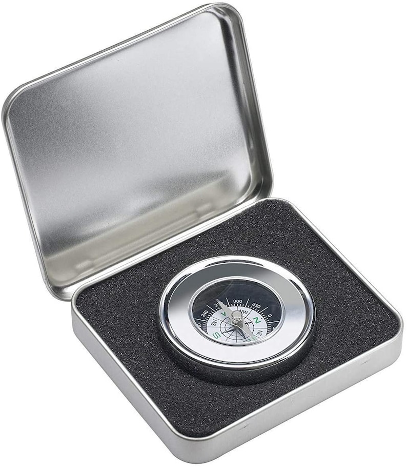 Puede incluir: Una br&uacute;jula plateada con una cara negra y marcas blancas. La br&uacute;jula est&aacute; en una caja de metal plateada con un forro de espuma negra.