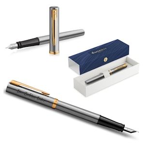 Könnte beinhalten: Ein silberner Füllfederhalter mit goldenen Akzenten, einer schwarzen Feder und dem Text "Dr. Sabine Dürr". Der Stift wird mit seiner Schachtel mit blau-weißem Design und einem zweiten Stift ohne Kappe präsentiert.