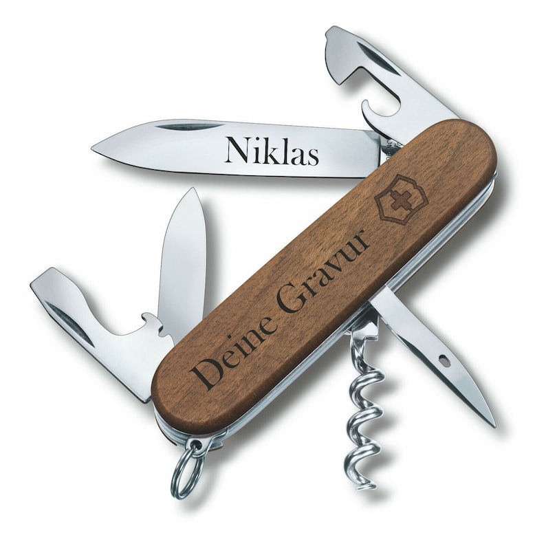 Puede incluir: Una navaja suiza con mango de madera y varios utensilios de acero inoxidable, incluyendo una hoja grabada con el nombre "Niklas". El mango tambi&eacute;n presenta el texto "Deine Gravur" y un logotipo de cruz.