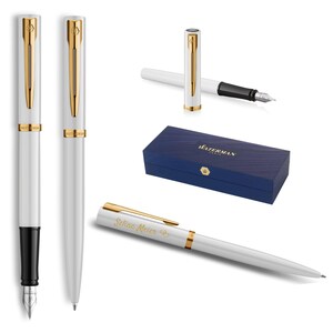 Könnte beinhalten: Weiße Füllfederhalter mit goldfarbenen Akzenten und schwarzen Federn werden gezeigt. Ein Stift ist geöffnet, ein anderer hat eine Kappe. Eine blaue Schachtel mit dem Markennamen "Waterman" ist ebenfalls abgebildet. Ein Stift ist mit dem Namen "Selina Meier" graviert.
