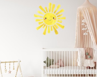 May include: A cheerful yellow sun wall decal with a smiling face and radiating rays. The watercolor design is applied to a white wall above a white crib, creating a bright and playful nursery theme.
