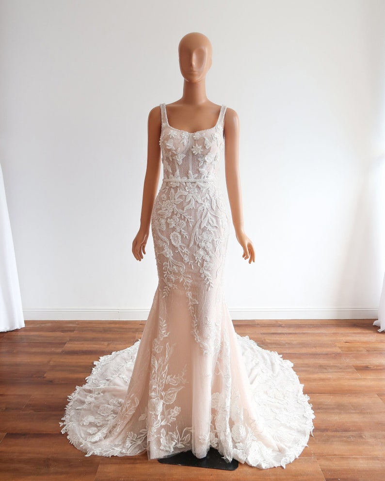 May include: A white and blush-colored wedding dress with a fitted bodice and a flowing skirt. The dress features delicate floral embroidery and a sweetheart neckline.