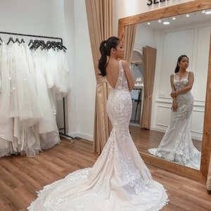 May include: A woman in a white wedding dress with a long train and intricate lace detailing. The dress has a low back and a fitted bodice. The woman is standing in a shop with a mirror behind her.