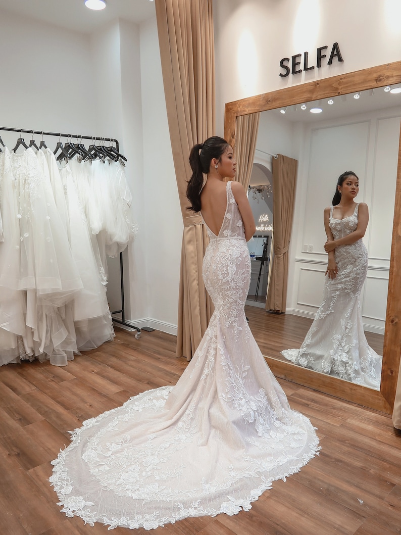 May include: A woman in a white wedding dress with a long train and intricate lace detailing. The dress has a low back and a fitted bodice. The woman is standing in a shop with a mirror behind her.