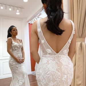 May include: A white wedding dress with a deep V-back and intricate floral lace detailing. The dress has a fitted bodice and a flowing skirt.