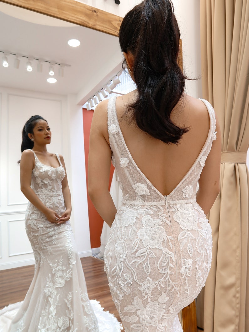 May include: A white wedding dress with a deep V-back and intricate floral lace detailing. The dress has a fitted bodice and a flowing skirt.