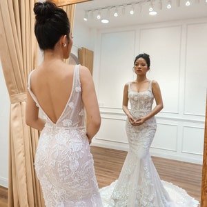 May include: A woman in a white, lace wedding dress with a fitted bodice and a flowing skirt. The dress features delicate floral embroidery and a low back.