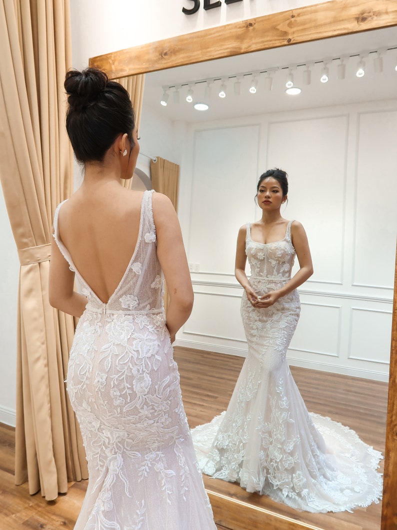 May include: A woman in a white, lace wedding dress with a fitted bodice and a flowing skirt. The dress features delicate floral embroidery and a low back.