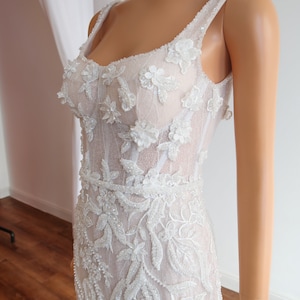 May include: A white wedding dress with a fitted bodice and a flowing skirt. The bodice is covered in intricate floral embroidery and beading. The skirt is made of a sheer fabric and has a train.