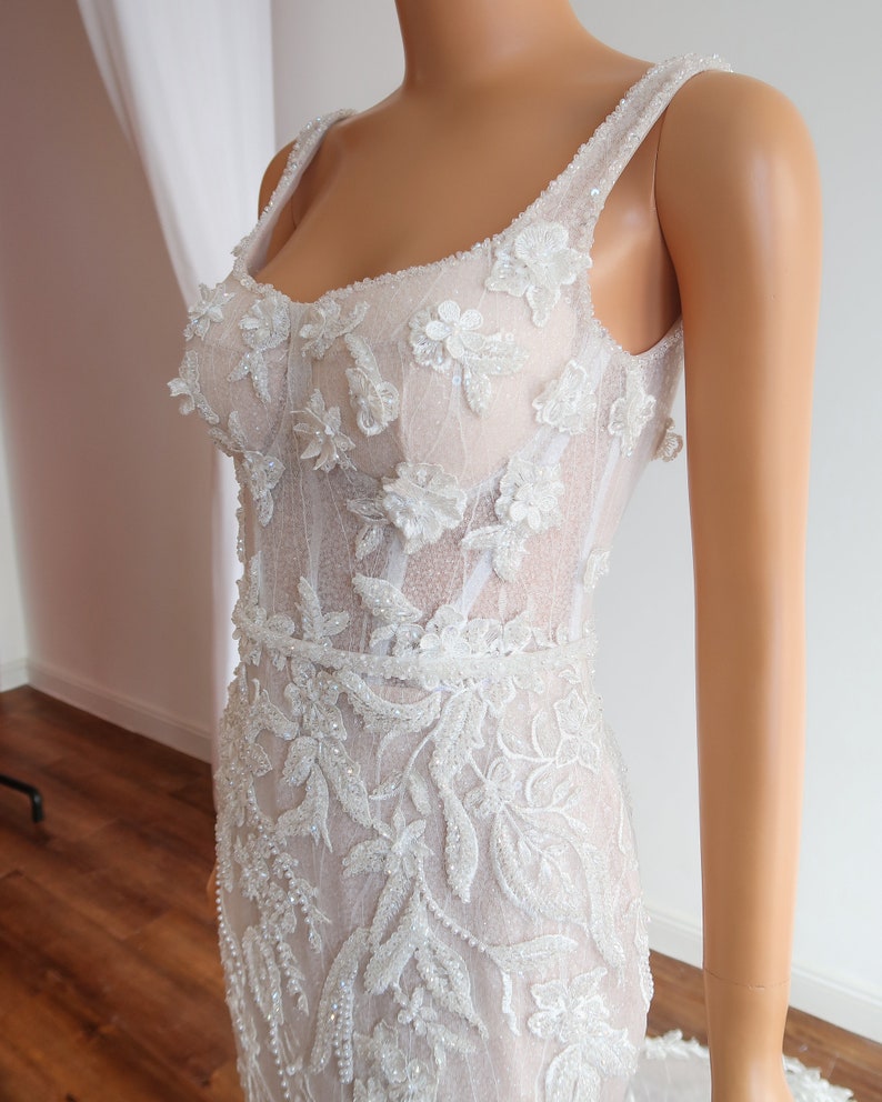 May include: A white wedding dress with a fitted bodice and a flowing skirt. The bodice is covered in intricate floral embroidery and beading. The skirt is made of a sheer fabric and has a train.
