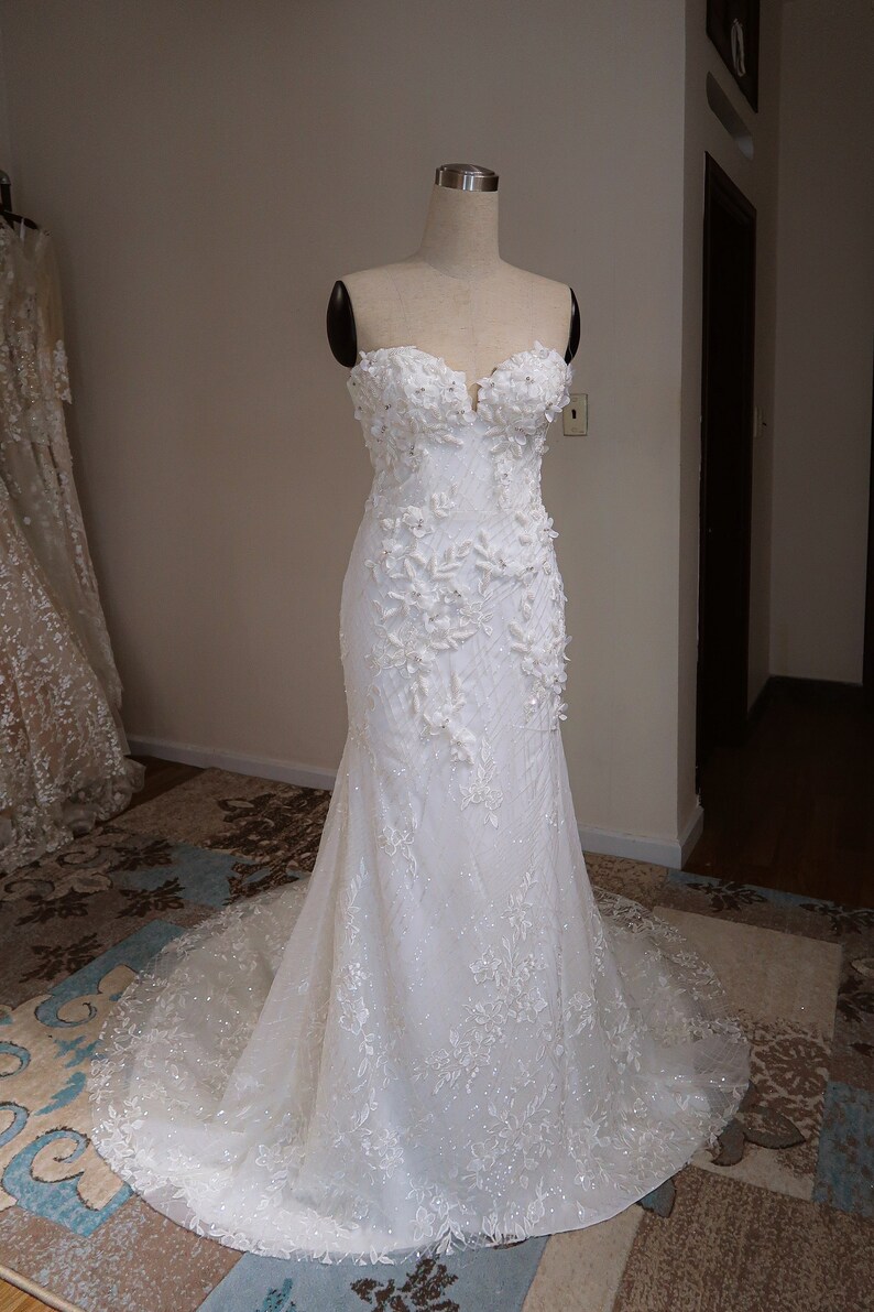 May include: A white strapless wedding dress with a fitted bodice and a flowing skirt. The dress is adorned with delicate floral appliques and sequins.