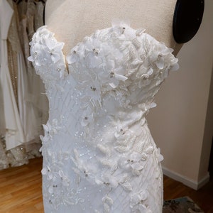 May include: A white strapless wedding dress with a fitted bodice and a flowing skirt. The bodice is adorned with delicate floral appliques and sequins.
