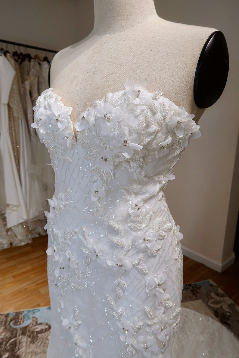 May include: A white strapless wedding dress with a fitted bodice and a flowing skirt. The bodice is adorned with delicate floral appliques and sequins.