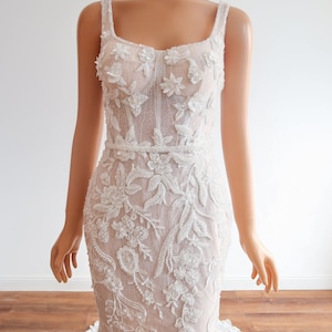 May include: A white wedding dress with a fitted bodice and a flowing skirt. The bodice is covered in delicate floral embroidery and sequins. The skirt is made of a sheer fabric with a subtle shimmer.
