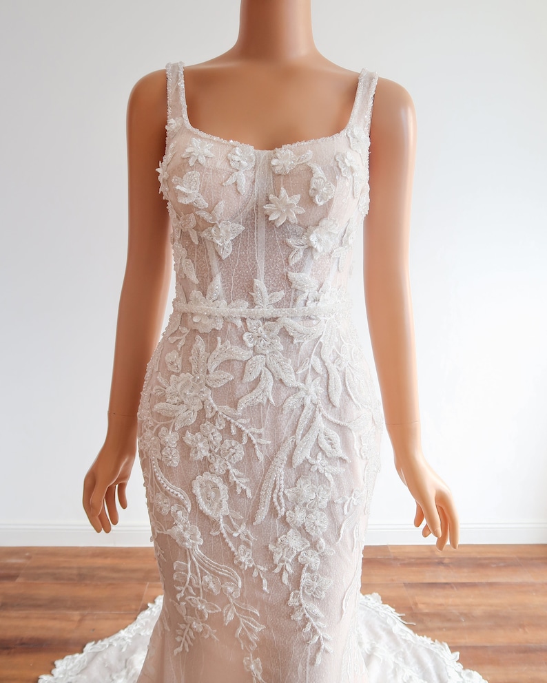 May include: A white wedding dress with a fitted bodice and a flowing skirt. The bodice is covered in delicate floral embroidery and sequins. The skirt is made of a sheer fabric with a subtle shimmer.
