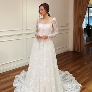 Pode incluir: Um vestido de noiva branco com mangas compridas e um corpete de renda. A saia é cheia e fluida, com um delicado padrão floral.