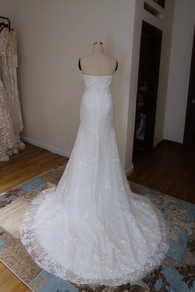 May include: A white strapless wedding dress with a fitted bodice and a flowing skirt. The dress is covered in delicate lace and sequins.